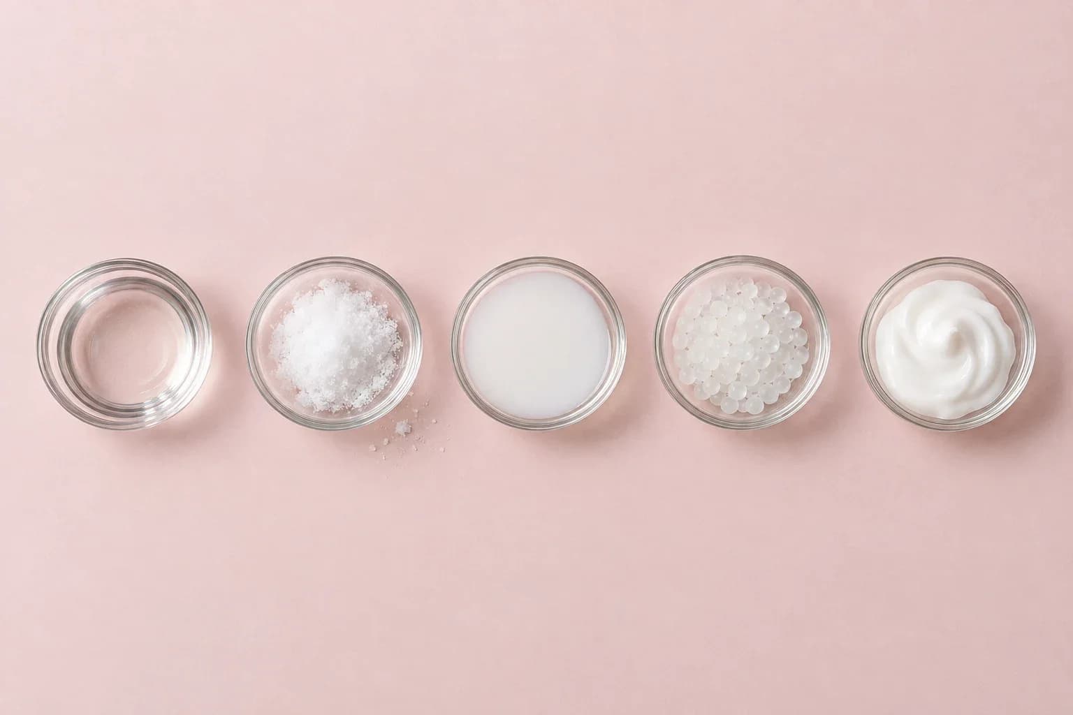 Five small glass dishes in a row, each holding a different raw material: clear liquid, powder, milky liquid, pellets, cream